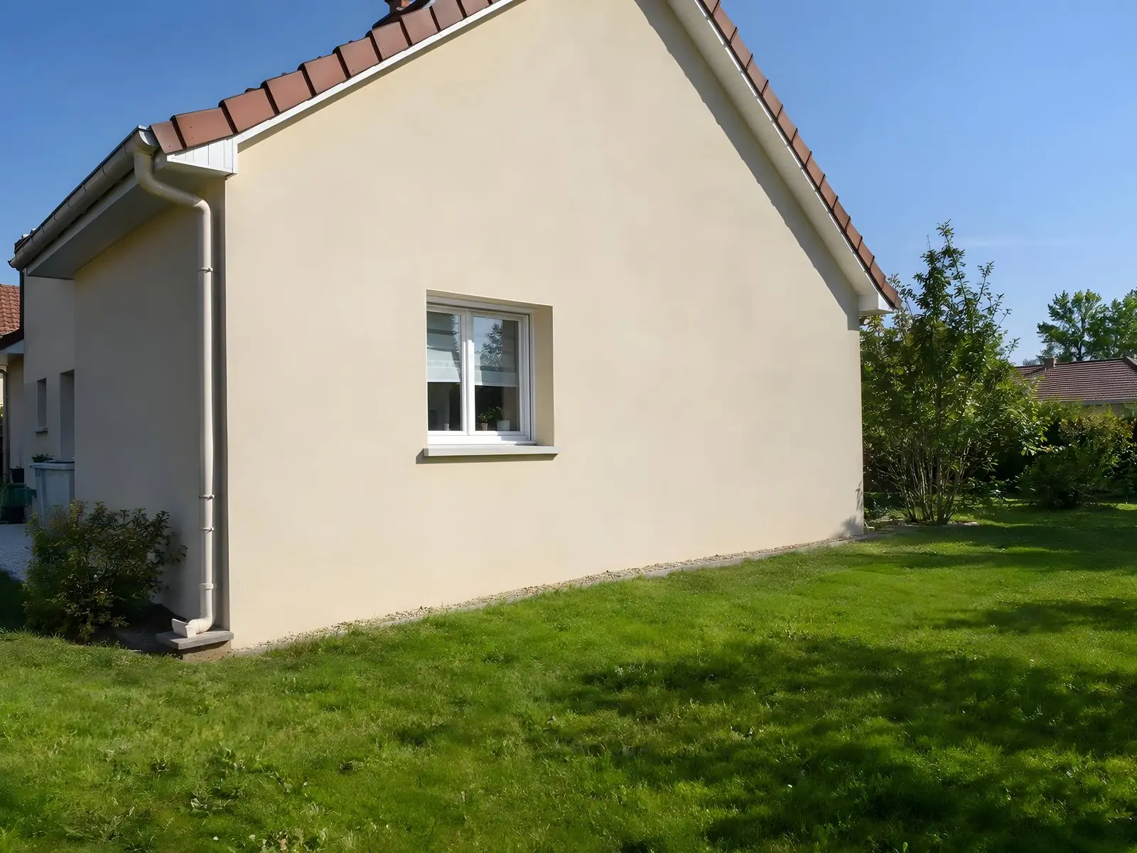 Pignon crépi beige d’un pavillon en plein soleil sous un toit de tuiles rouges, pelouse verte dégagée au pied du mur