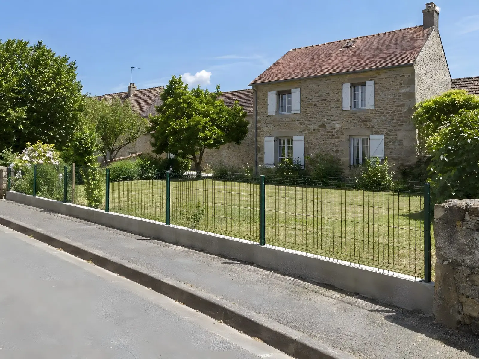 Terrain fauché court derrière un grillage vert bas, trottoir dégagé, maison en pierre aux volets clairs au fond