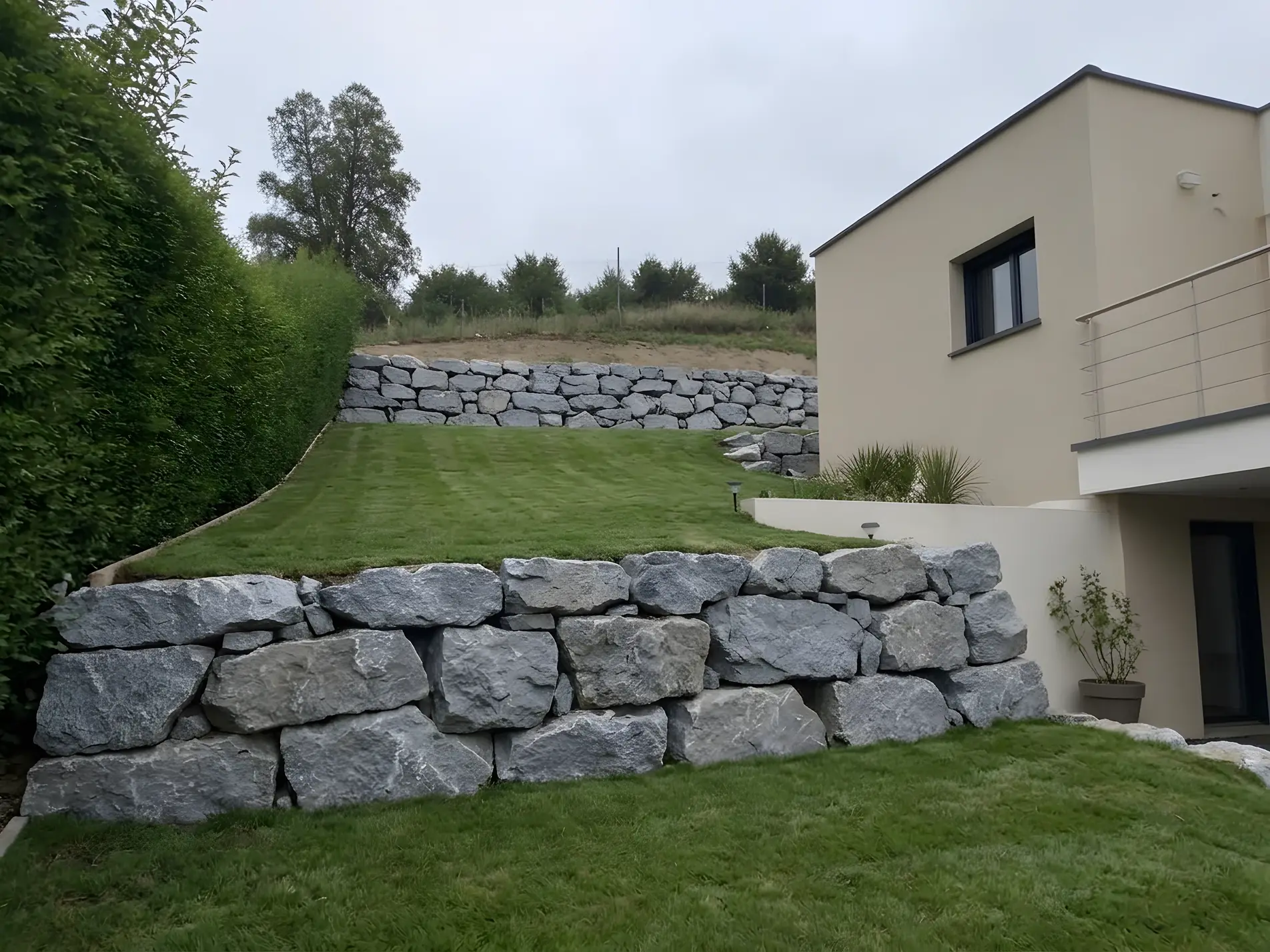 Terrain tenu net à côté d’une maison neuve : pelouse rase en terrasses, murets de gros blocs de pierre, haie taillée
